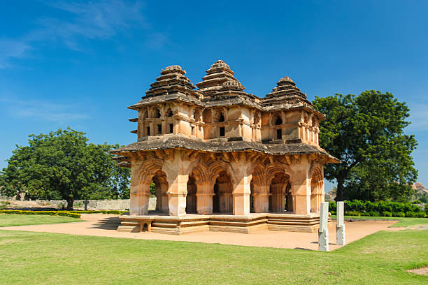 Ornate two-story pavilion featuring Islamic and Hindu architectural elements, known for its archways and lotus bud carvings. Constructed with a mix of stone and plaster, it features gracefully curved arches and domed structures on the upper level. Surrounded by lush greenery and a well-kept lawn, under a clear blue sky. The structure exudes historical grandeur, indicative of the Vijayanagara Empire s architectural style. The intricate details highlight the fusion of cultural influences in its design.