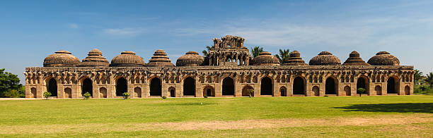 Symmetrical stone structure with a row of large arches and eleven intricately designed domes, showcasing Indo-Islamic architecture. Located in Hampi, India, these ancient elephant stables were used by the Vijayanagara Empire. The domes vary in shape, including circular and ribbed patterns, set against a backdrop of clear blue sky and palm trees. The foreground features a well-maintained grassy area, adding to the historical ambiance of the site.