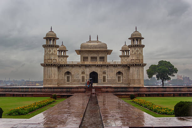 Mughal architecture featuring intricate pietra dura inlay work. The building, likely a tomb, has symmetrical design with prominent domes and minarets. Crafted from white and red sandstone, its facade displays detailed geometric and floral patterns. A pathway leads to the entrance, flanked by manicured lawns and gardens. Overcast skies create a dramatic backdrop, enhancing the structure s historical ambiance.