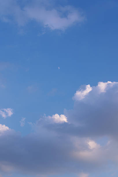 Dramatic sky with blue tones, fluffy white clouds, dark storm clouds, and a visible daytime moon. Peaceful yet moody natural sky background.
