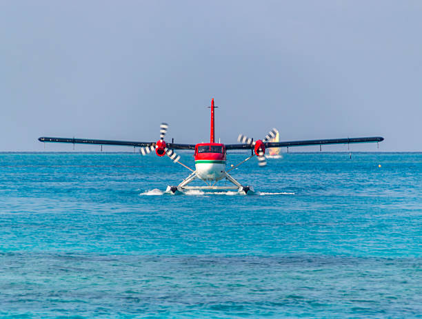 A vibrant seaplane with red, white, and black markings is positioned on the clear, turquoise water, seemingly preparing for takeoff. Its propellers are blurred, indicating motion. The calm ocean stretches out to a distant horizon under a bright, pale blue sky. The scene conveys a sense of adventure and travel in a stunning tropical location.