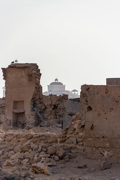 Ancient mud-brick ruins stand weathered and crumbling in the foreground, with a modern building featuring a large dome visible in the hazy distance, showcasing a blend of past and present architecture in the UAE.