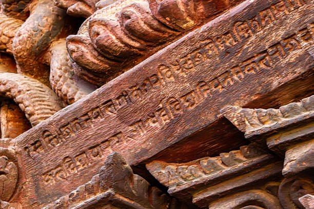 A close-up of an ancient wooden beam in Nepal, featuring intricate carvings and inscribed Sanskrit or Newari script, showing weathered textures and historical detail.