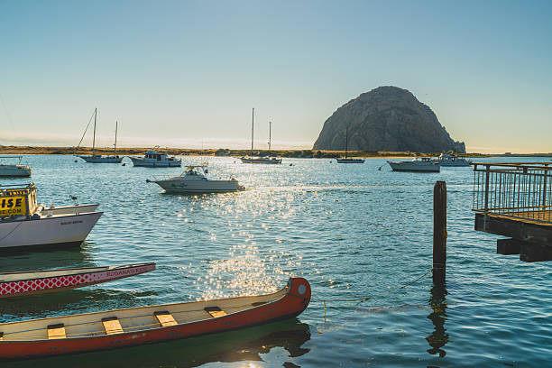 Morro Bay, California, USA. July 28, 2024. Boats float on shimmering water with Morro Rock in the background under a bright sky.