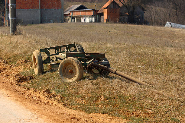 A single-axle green metal cart with large wheels lies unused on dry grassland near village houses, showing signs of rust and age in a quiet agricultural landscape