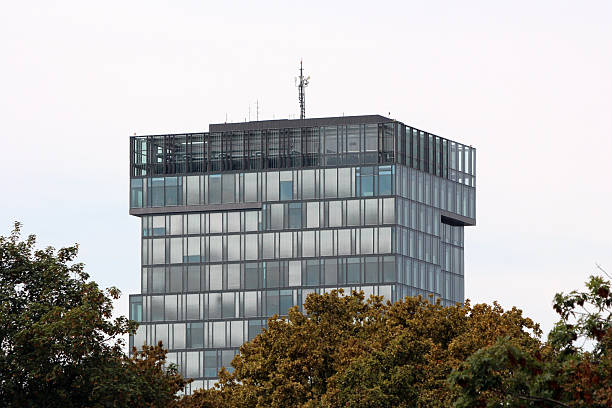 A multi-story modern office building with a façade of glass panels, a distinctive rectangular top, and rooftop antennas rises above tree foliage in a contemporary urban environment