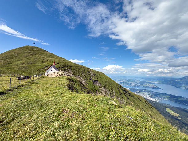 Small white chapel with red roof on grassy mountain ridge under blue sky. Scenic alpine landscape with rolling hills and distant lake view. Peaceful nature setting with panoramic mountain scenery.