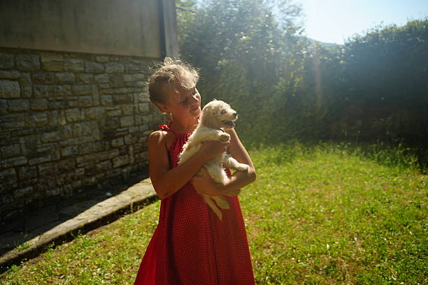 Young child in a red dress holds a small puppy in a sunny garden. The child smiles warmly, expressing joy and affection. Bright greenery and stone wall form the background setting.