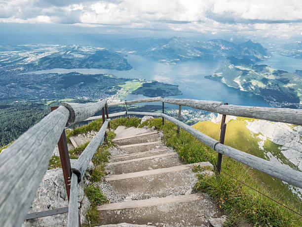 Wooden stairs with railing lead down a mountain path overlooking a large lake and valley. Scenic hiking trail with panoramic views of nature and distant towns. Outdoor adventure and landscape.