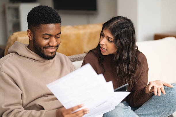 Couple discusses financial documents while sitting on a couch in a cozy living room during the afternoon, focusing on budgeting and planning for the future A man and a woman engage in a discussion about financial documents while seated in a comfortable living room, expressing various emotions during the conversation. tax planning discussion stock pictures, royalty-free photos  images
