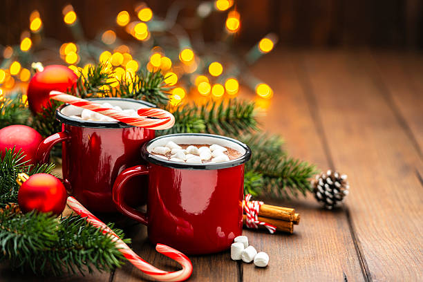 istock Two red mugs filled with hot chocolate and marshmallows on Christmas decorated table. Sweet food for winter or December Holidays. String lights, fir tree and candy canes 2228310676