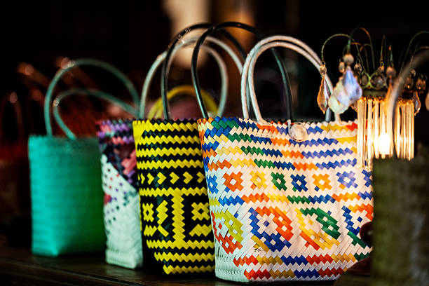 A vibrant row of handwoven bags and baskets, crafted from plastic ribbons or natural fibers, arranged on a dark surface in a market-like setting. Featuring bright colors and intricate patterns—including yellow, multicolored, and turquoise designs—the display highlights traditional craftsmanship and artisanal detail.