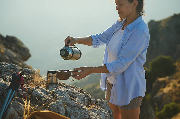 A woman pours coffee from a thermos into a cup while exploring a rocky terrain outdoors, surrounded by a scenic mountainous view under a tranquil sky.