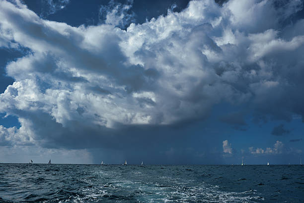 Sailing yachts scattered on the horizon under a dramatic, cloudy sky over open water. The sea shows dynamic waves and traces of a boat, while a storm builds in the distance. Captured before rainfall.