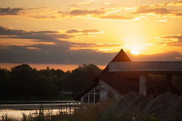 Golden sunlight casts a warm glow over a serene riverbank, highlighting a rustic building and trees while the sun sets on the horizon, creating a peaceful atmosphere.