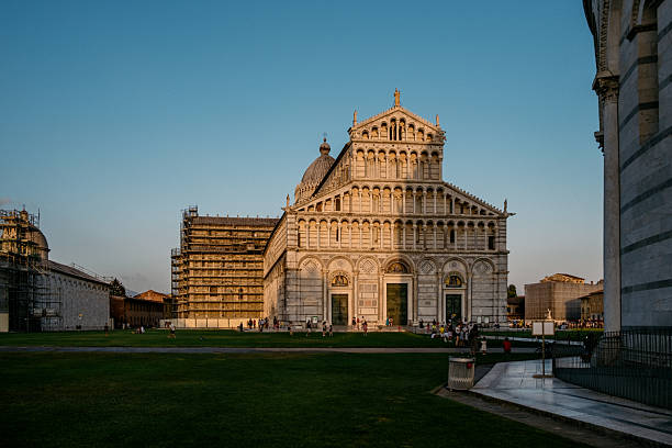 The iconic Cathedral of Pisa illuminated by golden sunset light at Piazza dei Miracoli, Italy, with tourists scattered across the green lawn.