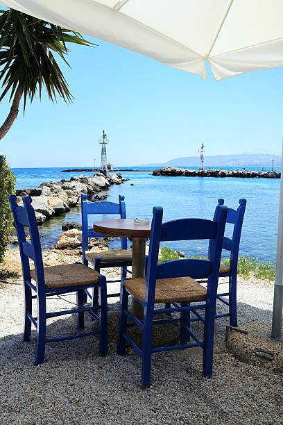 istock Traditional Greek taverna chairs and round table under a white umbrella facing the tranquil sea and lighthouse at Georgioupolis port, Crete. Ideal summer travel concept 2228348677
