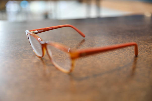 Selective Focus On Stylish Red-Temple Eyeglasses Resting On A Wooden Table. Blurred Background Highlights The Smooth Surface And Casual Ambiance, Suggesting Everyday Use Or Pause From Reading.