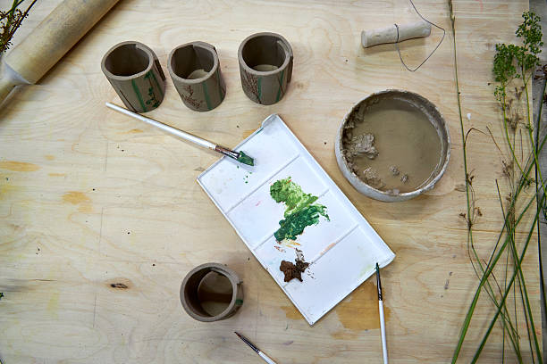 Hand-painted clay napkin rings with botanical imprints in progress. Brushes, paint palette, and slip bowl on a wooden table highlight the artistic stage of decorating handmade ceramic tableware