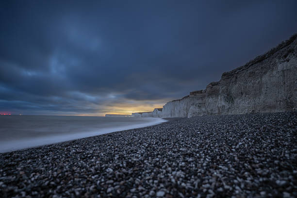 A stunning long-exposure night shot of Beachy Head and the iconic Seven Sisters chalk cliffs along the southern English coast. The image captures the ethereal glow of the night sky above the dramatic white cliffs, blending natural grandeur with a serene, moody atmosphere.
