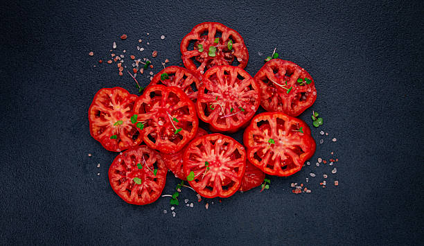 istock Freshly sliced ripe tomatoes arranged on a textured dark surface with herbs and salt scattered around 2228367303