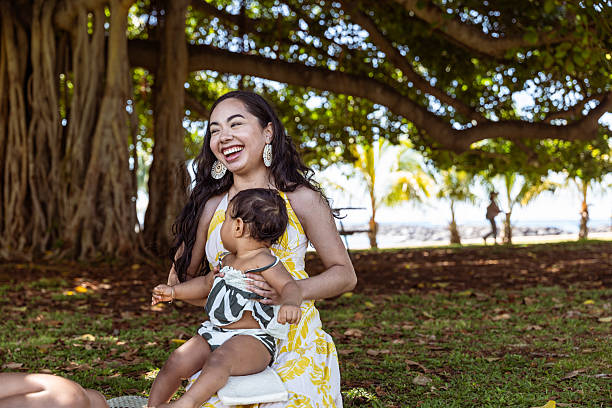 A smiling Pacific Islander woman holds her baby niece on her lap while sitting under a banyan tree in Hawaii.