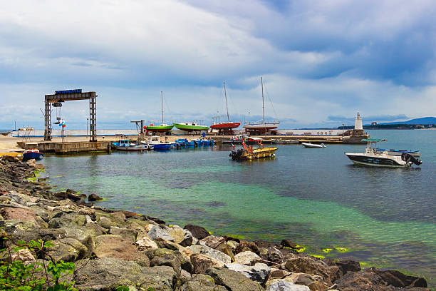 Primorsko, Bulgaria - May 26, 2021: Scenic view towards the whitewashed lighthouse at the port with sailboat and fishing boats in the shallow water in front