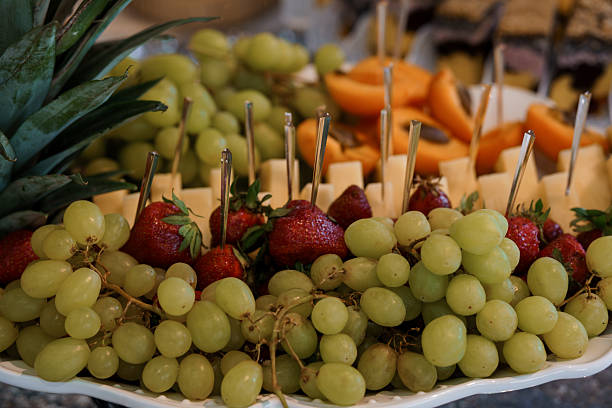 A close-up shot showcases a beautifully arranged fruit platter with fresh green grapes, ripe strawberries, a slice of pineapple, cantaloupe, and cheese cubes skewered. perfect for healthy eating or party catering