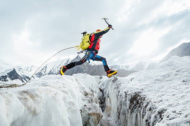 Climber in vibrant gear leaps over crevasse on icy glacier during Lenin peak ascent under clear sky with snow-covered peaks in background. Extreme active people, high-altitude mountaineering concept.