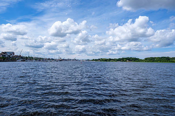 Scenic view of a harbor with sailboats and a calm river under a vibrant blue sky filled with fluffy cumulus clouds. The waterfront is lined with trees and buildings, while industrial cranes are visible in the distance.