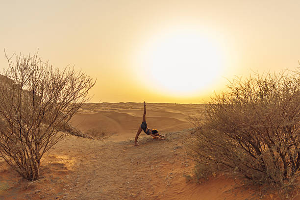 A person performs yoga in a tranquil desert during sunset. The golden light complements the serene environment, symbolizing relaxation and balance. The sandy dunes and sparse desert plants add to the sense of peace, while the harmonious pose portrays a connection to nature and mindfulness.