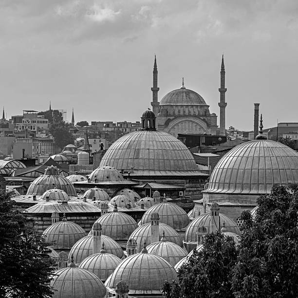 A layered view of Ottoman architecture captured from the courtyard of Suleymaniye Mosque in Istanbul. The image showcases a cluster of historic domes in the foreground, gradually revealing the cityscape of Eminonu and the prominent Rüstem Pasha Mosque in the distance