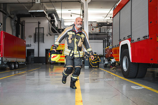 Wide-angle shot of mid-aged firefighter in reflective yellow-black turnout gear sprinting through airport fire station. Bright industrial lighting illuminates red fire trucks and concrete floor.