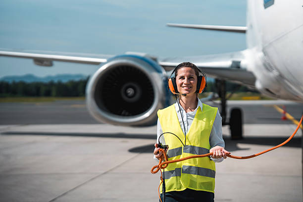 A mid adult woman of Caucasian descent holds a corded headset beside a stationary cargo plane while dressed in standard ramp safety gear during active ground support duties.