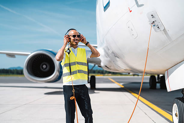 Silhouette shot of a young adult male passenger standing and listening to music with headphones. He is at the gate waiting for the flight. At the background there is a airplane parked ready to board.