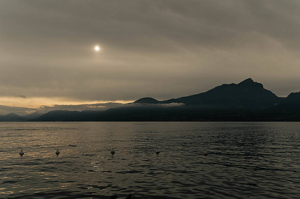 Lake Garda is captured in late afternoon light with a cloudy sky. The sun appears as a soft glowing disk behind the clouds, creating a calm and moody atmosphere.