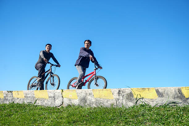 Two Young Men Ride Bicycles On A Sunny Day, Enjoying An Outdoor Adventure Under A Clear Blue Sky