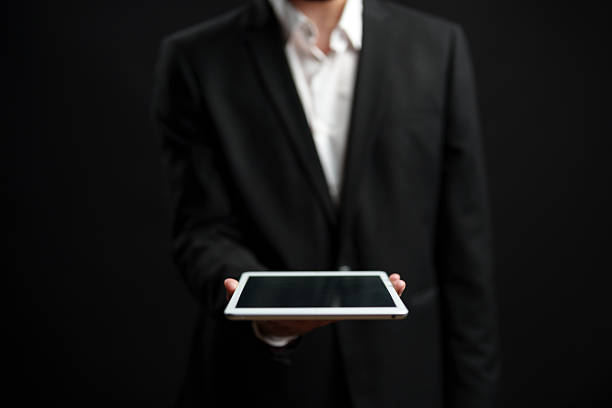 A business professional in a suit holds a tablet in front of a dark background, highlighting the importance of technology in modern communication and work.