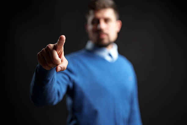 A man wearing a blue sweater gestures with his finger, creating a sense of emphasis. The dark background enhances the focus on his action and expression.