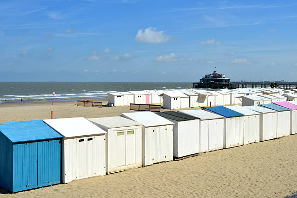 Blankenberge, West-Flanders, Belgium- August 3, 2025: In the background, a Belle Époque-style restaurant building at the end of the pier. The  Blankenberge landmark. Beach huts in blue, white, and pink, arranged in a row on the sand, create a beautiful, coincidental color composition.