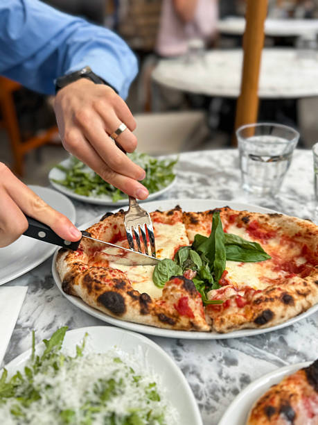 Stock photo shows a person is slicing and cutting up a fresh Margherita pizza topped with vibrant basil using a knife and fork at an outdoor pizzeria table. Sunlit, casual dining captures the authentic Italian experience, highlighting fresh ingredients, golden crust, melted cheese