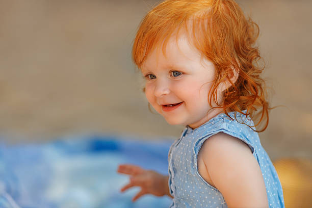 Cheerful red-haired toddler girl in a blue dress smiling and playing with her hands outdoors on a sunny summer day. Bright natural light, warm tones, and joyful expression .