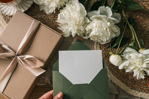 istock Green envelope with blank white card held by hand, next to kraft gift box with satin ribbon and white peonies on natural textured background. Elegant flat lay scene for celebration or invitation 2228438821
