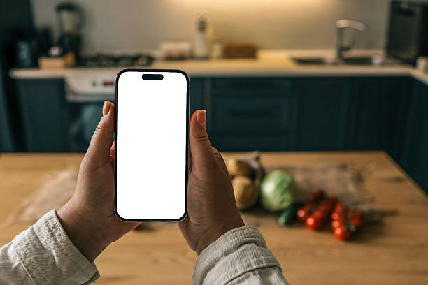 istock Person holding a smartphone with blank white screen over a rustic kitchen table filled with fresh vegetables: potatoes, tomatoes, cabbage, cucumber. Mockup concept with natural lighting 2228438950