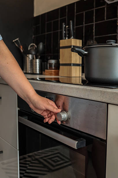 istock Woman adjusting oven temperature using a silver control knob in a modern kitchen with black tiles, utensils, and a pot on the stovetop 2228439533