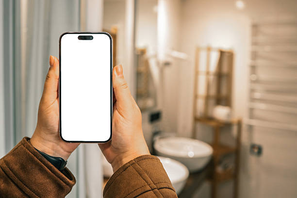 istock Person holding a smartphone with blank white screen in modern bathroom interior with sinks, mirror, and wooden shelf blurred in the background 2228439690