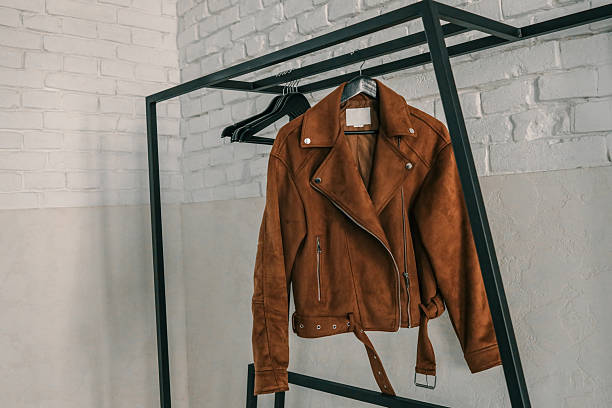 Brown suede jacket hanging on a black minimalist metal rack in a bright room with white brick walls and a mirror in the background.