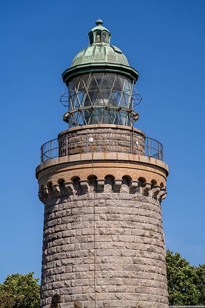 istock Historic stone lighthouse with a green dome under a clear blue sky, surrounded by trees. 2228441750