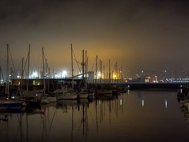 Foggy evening scene of a quiet urban marina where sailboats and yachts are moored, their silhouettes and harbor lights reflecting softly on still water.