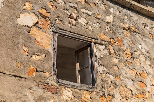 An abandoned structure features a weathered stone wall and a simple wooden window. Sunlight casts warm tones on the rustic texture, highlighting the village's charm